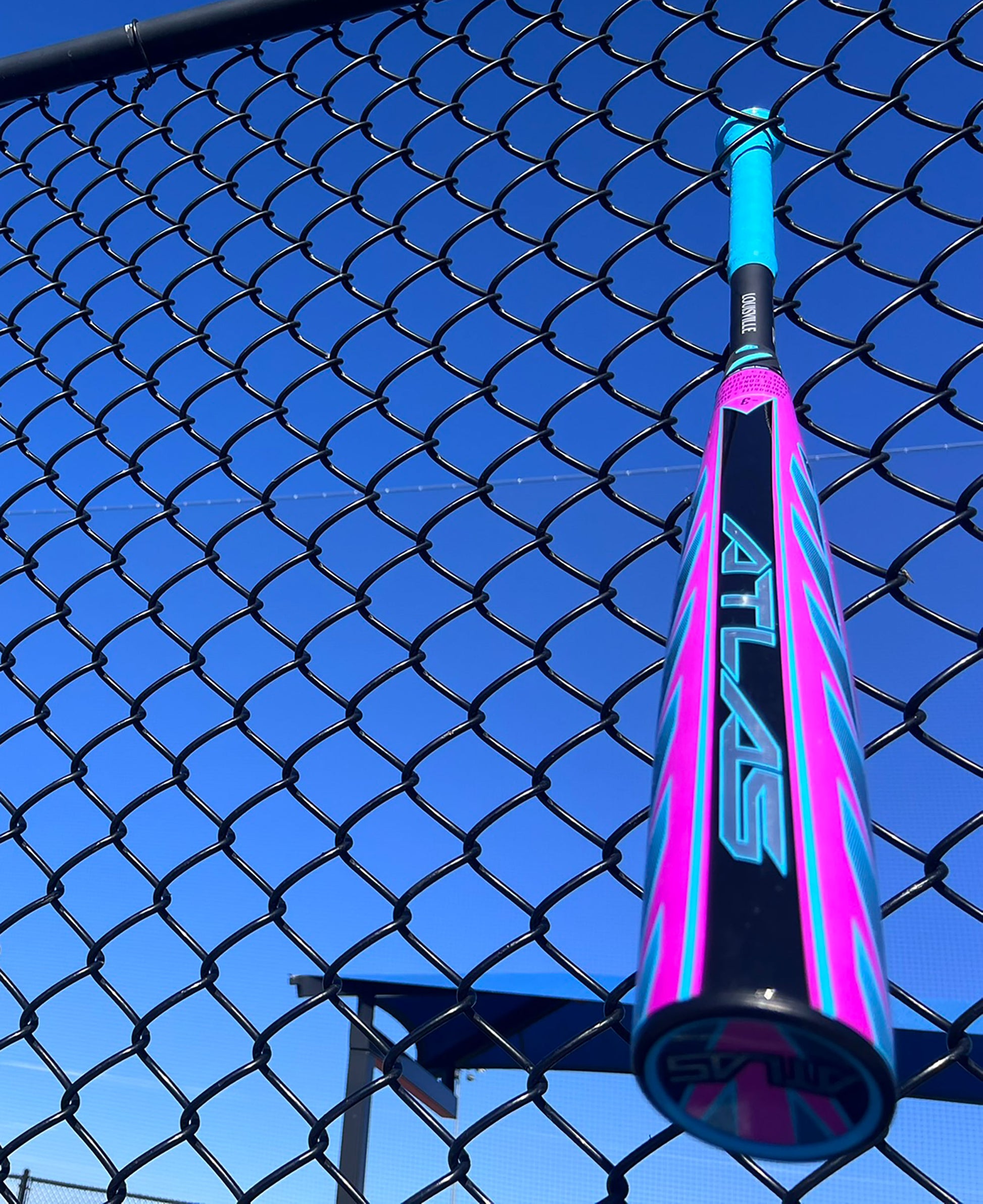A blue and pink 2026 Louisville Slugger Atlas Hybrid (-3) BBCOR Baseball Bat (WBL4114010) from Louisville Slugger rests between the links of a black chain-link fence beneath a clear blue sky.