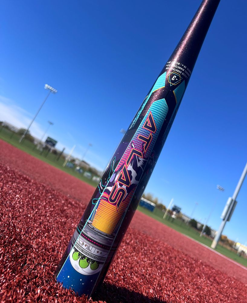 The 2026 Louisville Slugger Atlas A51 (-3) BBCOR Baseball Bat (WBL4180010) stands upright on red turf, showcasing its vibrant, space-themed design under a blue sky and bright stadium lights.