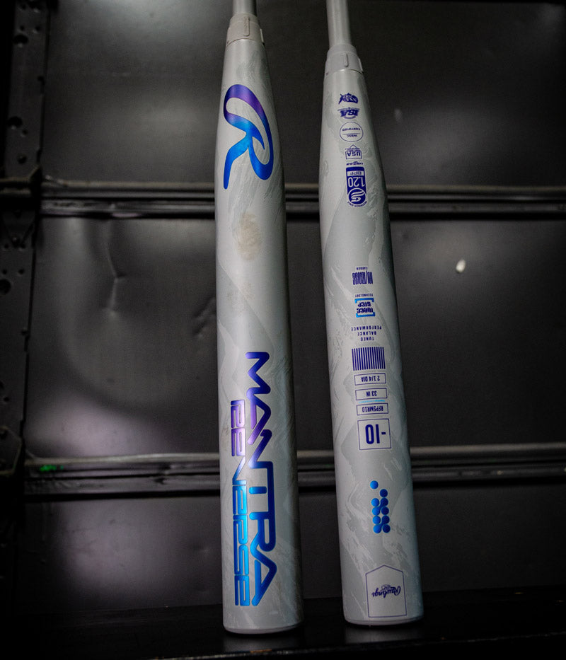 Two 2025 Rawlings Mantra Reverse (-10) fastpitch softball bats, white with blue MANTRA graphics and certification symbols, stand upright against a dark industrial background, highlighting their advanced Three-Step Technology.