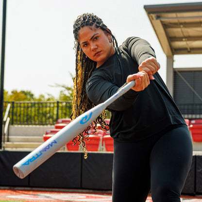 A woman with braided hair swings a 2025 Rawlings Mantra Reverse (-10) Fastpitch Softball Bat (RFP5MR10) on a field, dressed in black athletic wear, with empty red seats and a shaded structure visible in the background.