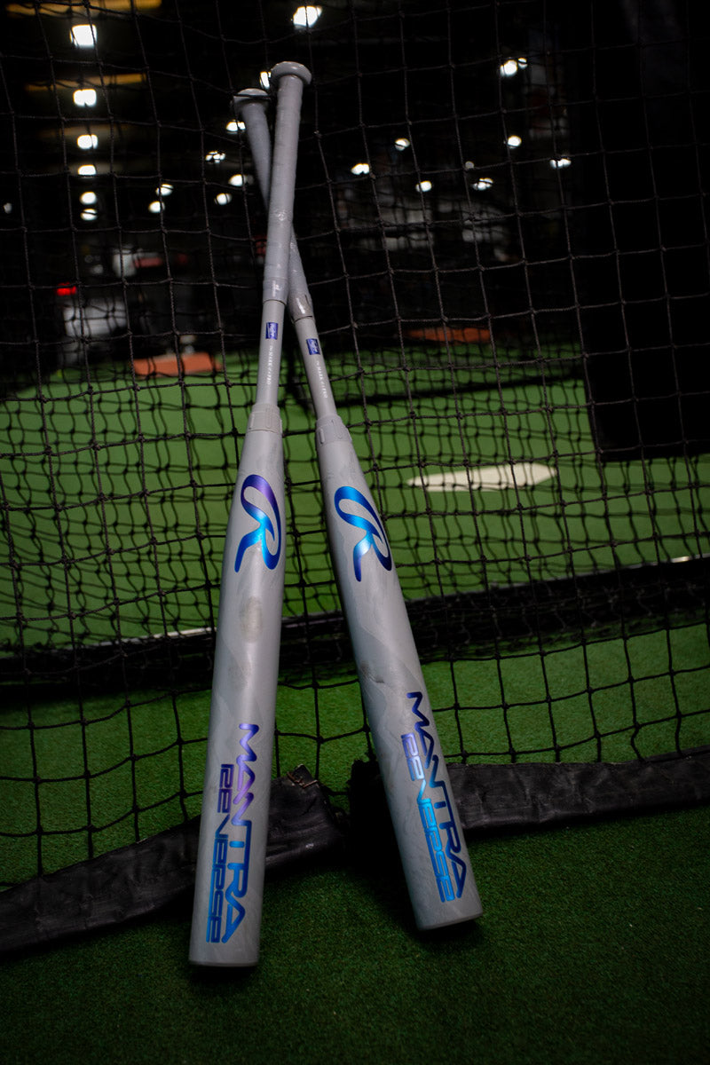 Two gray 2025 Rawlings Mantra Reverse (-10) Fastpitch Softball Bats are crossed and leaning against a black net in an indoor batting cage on green turf, featuring blue and purple logos and Three-Step Technology.