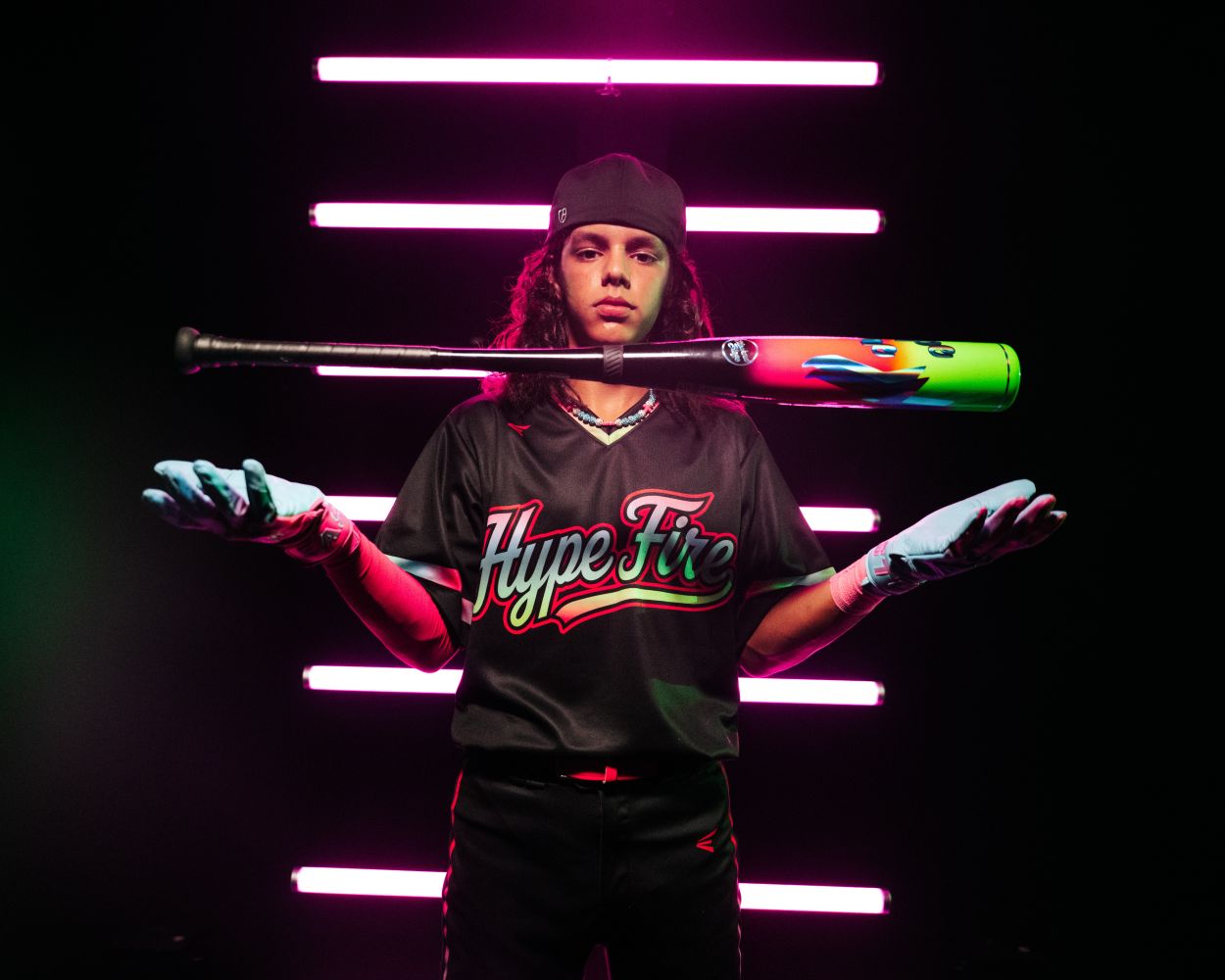 Wearing a Hype Fire jersey, gloves, and a backward cap, a person stands with the 2027 Easton Hype Fire (-10) 2 3/4″ USSSA Baseball Bat (EUT7HYP10) from Easton floating between their hands, set against a backdrop of pink neon lights.