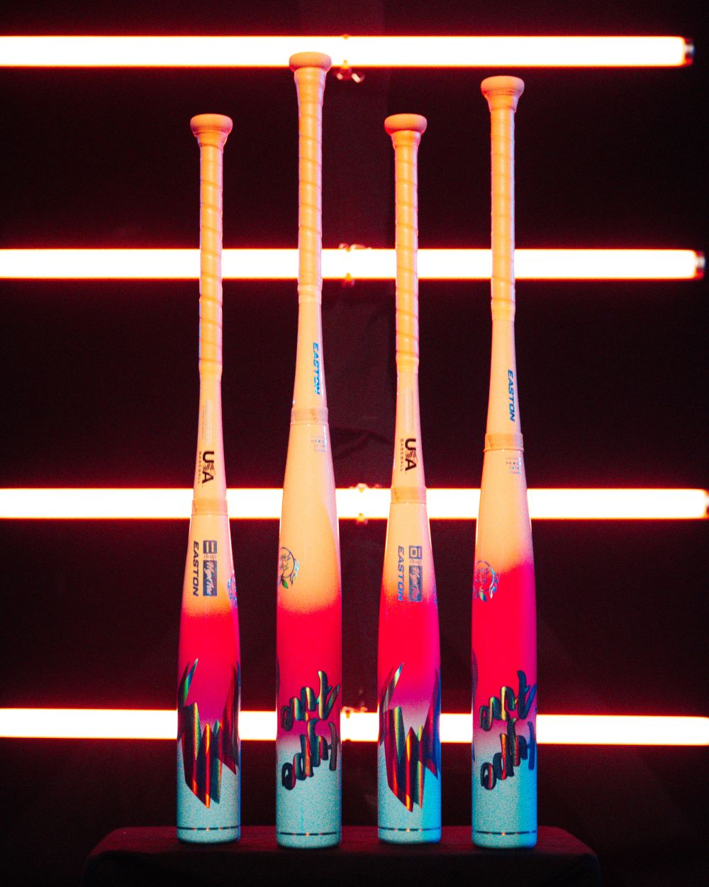 Four vibrant Easton Hype Fire (-11) 2 5/8″ USA Baseball Bats (EUS7HYP11) stand upright against a black background, illuminated by horizontal neon orange lights. Featuring advanced 3D Ropecoil Composite technology and the innovative ConneXion Max connection system, these Easton bats deliver both striking visuals with their gradient red, white, and blue designs and exceptional performance.