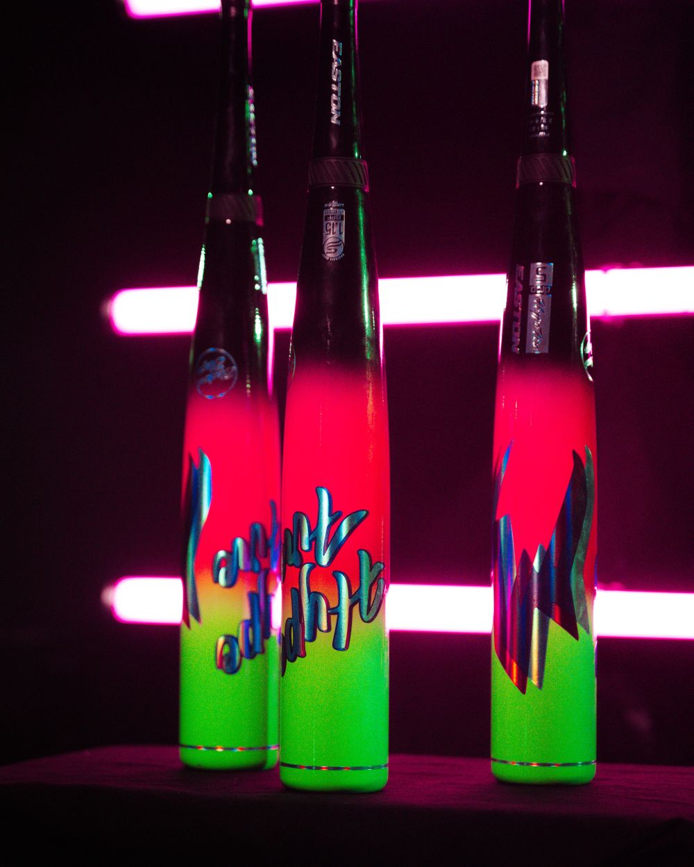 Three neon-colored baseball bats—including the powerful 2027 Easton Hype Fire (-10) 2 3/4″ USSSA Baseball Bat: EUT7HYP10 by Easton with its advanced TCT Barrel—stand upright against a black background with pink horizontal lights, each showcasing vibrant red, green, and blue designs.