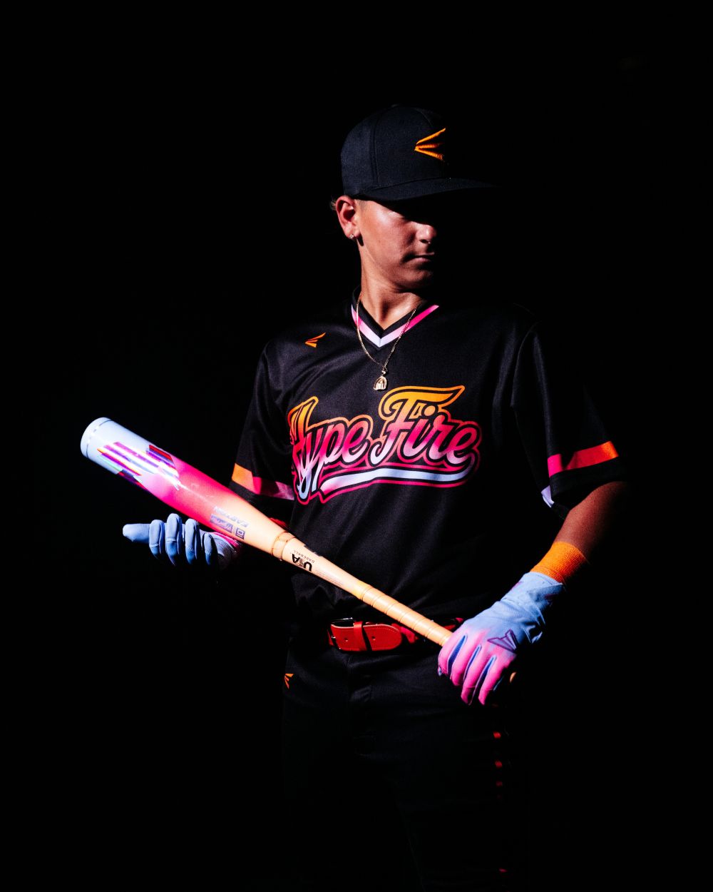 A baseball player wearing a black Hype Fire jersey, black cap, and blue gloves holds the 2027 Easton Hype Fire (-11) 2 5/8″ USA Baseball Bat (EUS7HYP11) by Easton, showcasing its signature pink and white design with advanced 3D Ropecoil Composite technology, against a dark background.