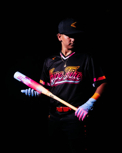 A baseball player wearing a black Hype Fire jersey, black cap, and blue gloves holds the 2027 Easton Hype Fire (-11) 2 5/8″ USA Baseball Bat (EUS7HYP11) by Easton, showcasing its signature pink and white design with advanced 3D Ropecoil Composite technology, against a dark background.