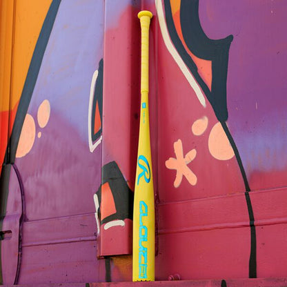 A vivid yellow 2026 Rawlings Clout AI (-3) BBCOR Baseball Bat (RBB6C3) with blue lettering stands against a purple and orange graffiti wall.