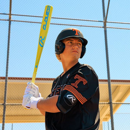 A baseball player in a black uniform and helmet prepares to hit with a 2026 Rawlings Clout AI (-3) BBCOR Baseball Bat (RBB6C3), standing by a chain-link fence under a blue sky.