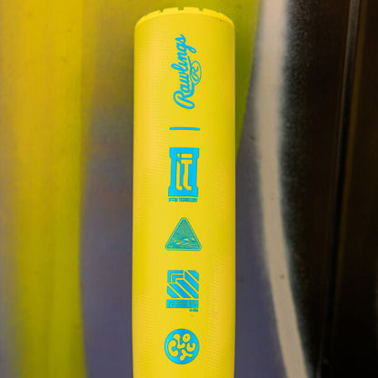 A close-up of the 2026 Rawlings Clout AI (-3) BBCOR Baseball Bat (RBB6C3) in bright yellow, featuring blue icons and the Rawlings logo, set against a background with vertical yellow, brown, and black streaks.