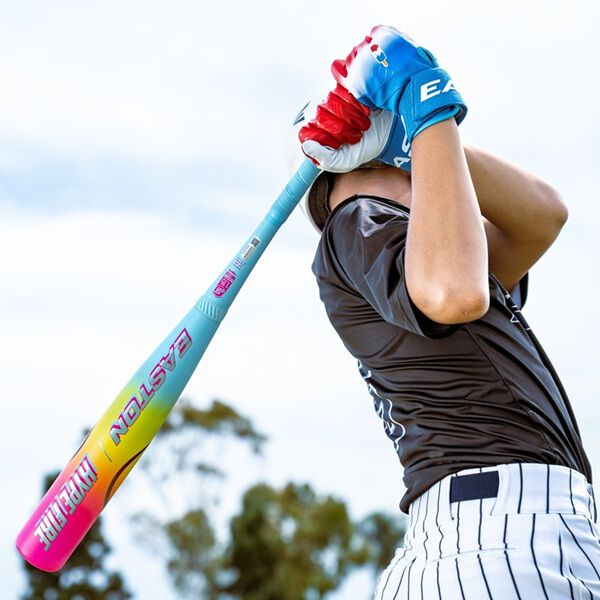 A baseball player in a dark jersey swings the 2026 Easton Hype Fire (-5) 2 3/4" USSSA Baseball Bat (EUT6HYP5) by Easton, wearing red and blue gloves, with trees and a cloudy sky in the background.