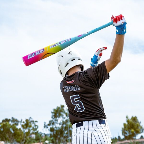 A baseball player in a white helmet, black jersey with number 5, and striped pants readies to swing the 2026 Easton Hype Fire (-5) USSSA Baseball Bat by Easton. Trees and a blue sky provide the backdrop.