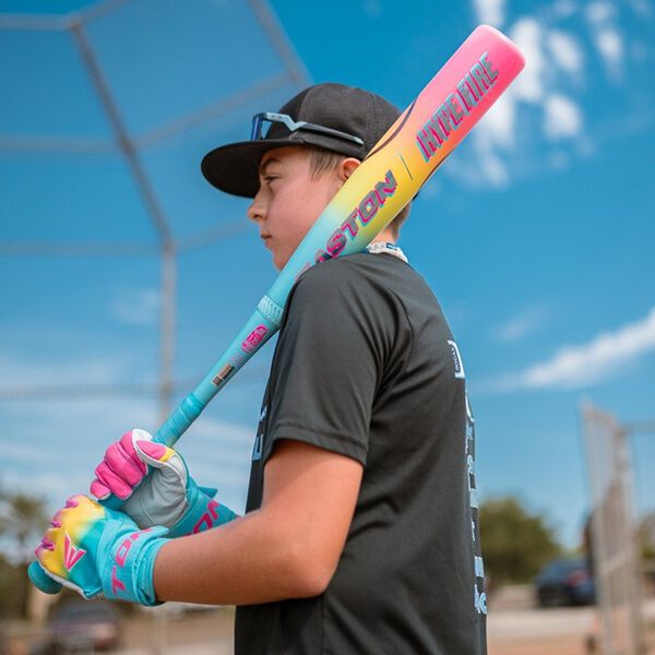 A young baseball player in a black shirt and cap holds the 2026 Easton Hype Fire (-5) 2 3/4" USSSA Baseball Bat: EUT6HYP5 over their shoulder while wearing gloves on a baseball field under a blue sky.