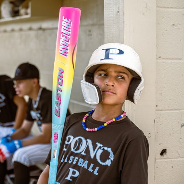 A young baseball player in a helmet and beaded necklace leans against a wall, holding the 2026 Easton Hype Fire (-5) 2 3/4" USSSA Baseball Bat (EUT6HYP5) by Easton. He wears a PONO BASEBALL shirt; two teammates sit on a bench in the background.