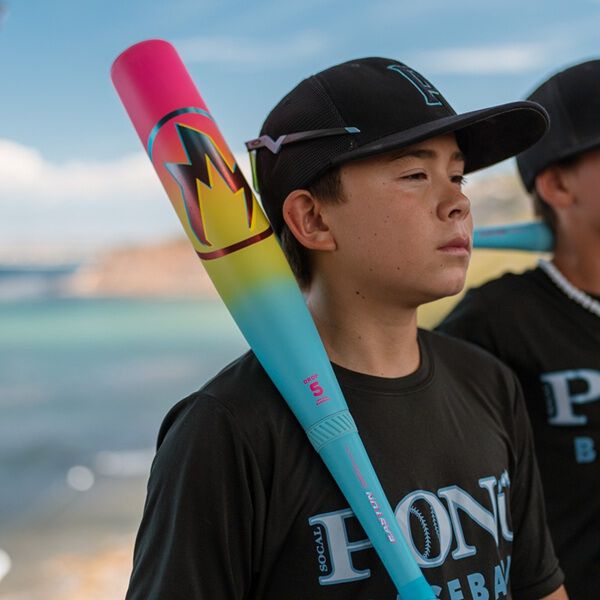 A boy in a black baseball cap and shirt holds the 2026 Easton Hype Fire (-5) 2 3/4" USSSA Baseball Bat (EUT6HYP5) over his shoulder, standing outdoors near water with another player visible in the background.
