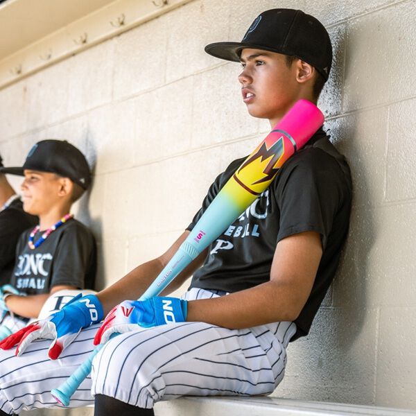 Two boys in baseball uniforms sit in a dugout. The boy in front grips an Easton 2026 Hype Fire (-5) 2 3/4" USSSA Baseball Bat and wears a black cap, black shirt, striped pants, and blue batting gloves. The other boy sits next to him.