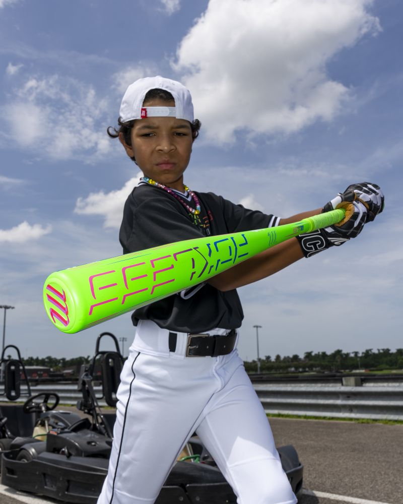 A young baseball player in uniform grips the vibrant green Marucci 2027 RCKLESS Gen 2 USA Baseball Bat, ready to swing. He sports a backwards cap and colorful necklaces, with go-karts and a cloudy sky visible in the background.