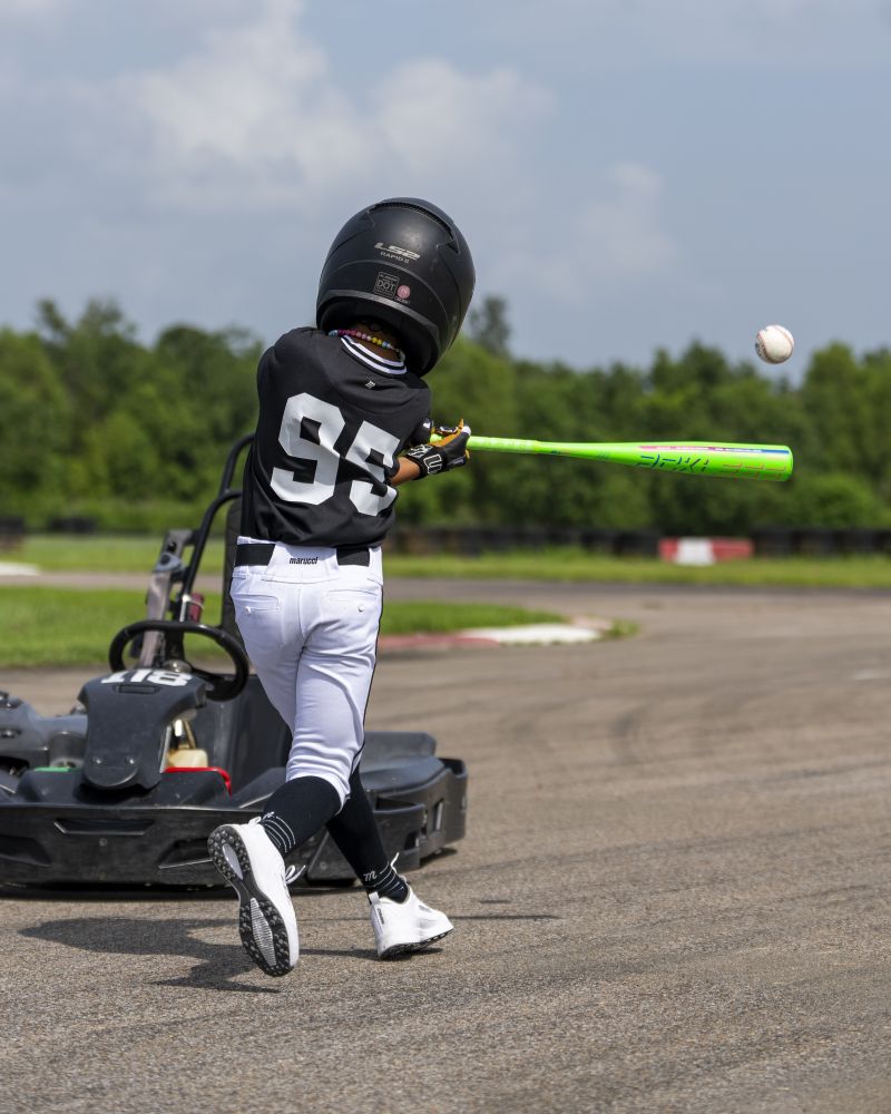Wearing a black helmet and a jersey with the number 95, a child stands near a go-kart on a track and swings the Marucci 2027 RCKLESS Gen 2 USA Baseball Bat at an incoming pitch. Trees and a blue sky provide the backdrop.