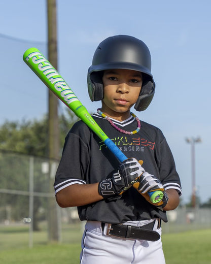 A young baseball player wearing a helmet and black uniform holds a neon green Marucci 2027 RCKLESS Gen 2 Hybrid (-8) 2 5/8″ USA Baseball Bat (MSBRH28U), which features Ring-Free Barrel Construction, standing on a field with a focused expression as trees and a fence appear in the background.