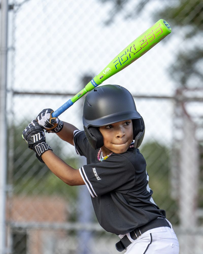 A young baseball player wearing a black helmet and uniform prepares to swing the Marucci 2027 RCKLESS Gen 2 Composite (-10) 2 5/8″ USA Baseball Bat (MSBRC210U) with maximum barrel flex during a game, standing ready at home plate with a focused expression.