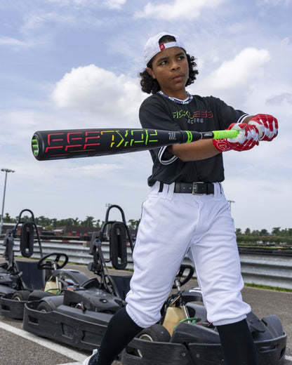 A young baseball player in uniform holds the Marucci 2027 RCKLESS Gen 2 Composite (-5) 2 3/4″ USSSA Baseball Bat (MSBRC25), featuring an ergonomic hand taper, while standing near parked go-karts outdoors under a partly cloudy sky.