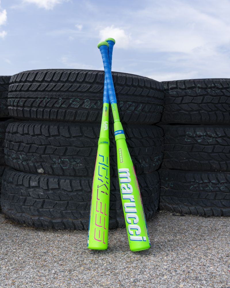 Two 2027 Marucci RCKLESS Gen 2 Composite (-10) USA Baseball Bats, each featuring neon green barrels and blue handles and engineered for maximum barrel flex, lean against a stack of worn black tires on an outdoor gravel surface, with a partly cloudy sky in the background.