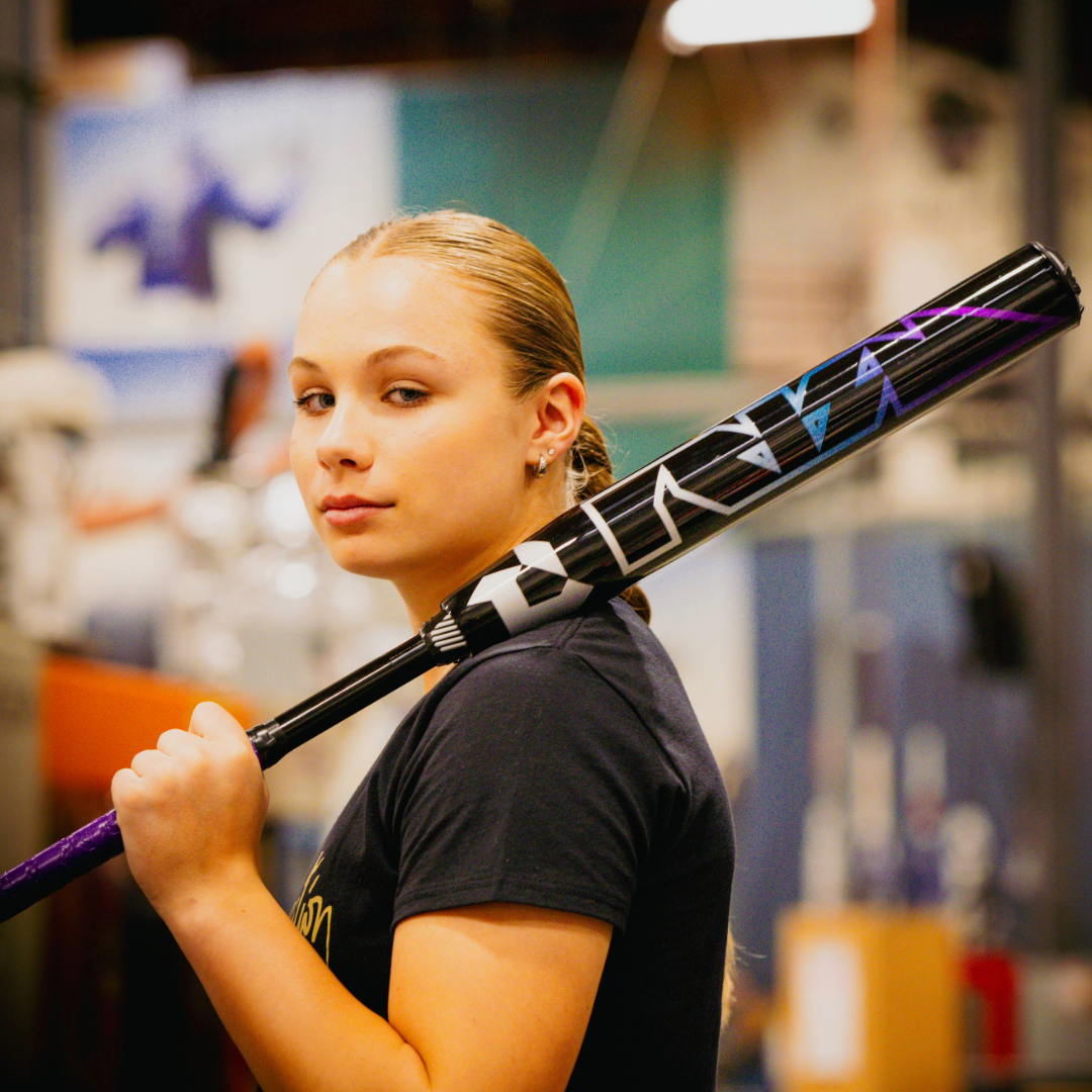 A young woman with blonde hair pulled back holds the 2026 DeMarini Prism+ (-11) FE Fastpitch Softball Bat (WBD2636010) over her shoulder, gazing confidently at the camera in an indoor setting. She wears a dark t-shirt.
