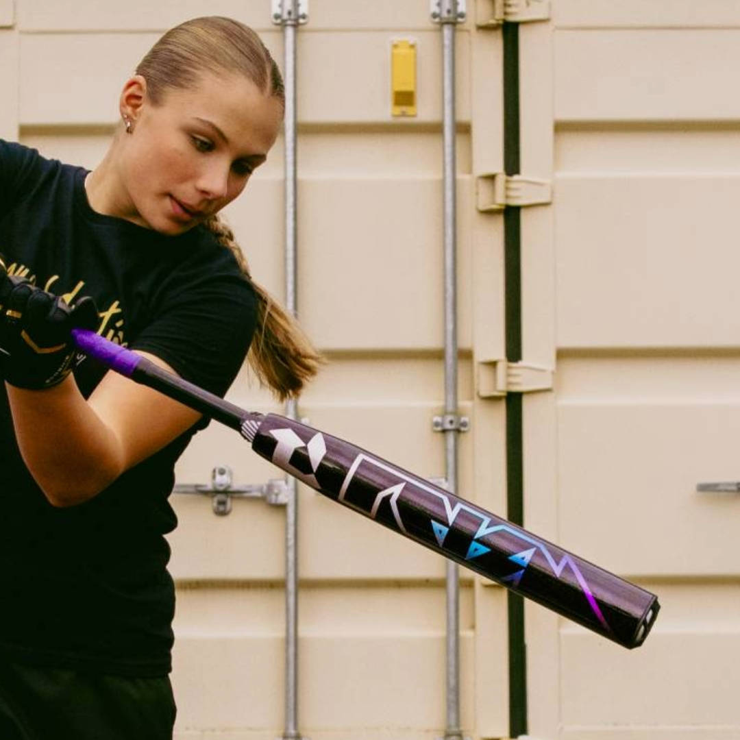 A young girl in a black shirt and gloves readies to swing a 2026 DeMarini Prism+ (-11) Fastpitch Softball Bat (WBD2636010), with a beige metal door featuring vertical lines in the background.