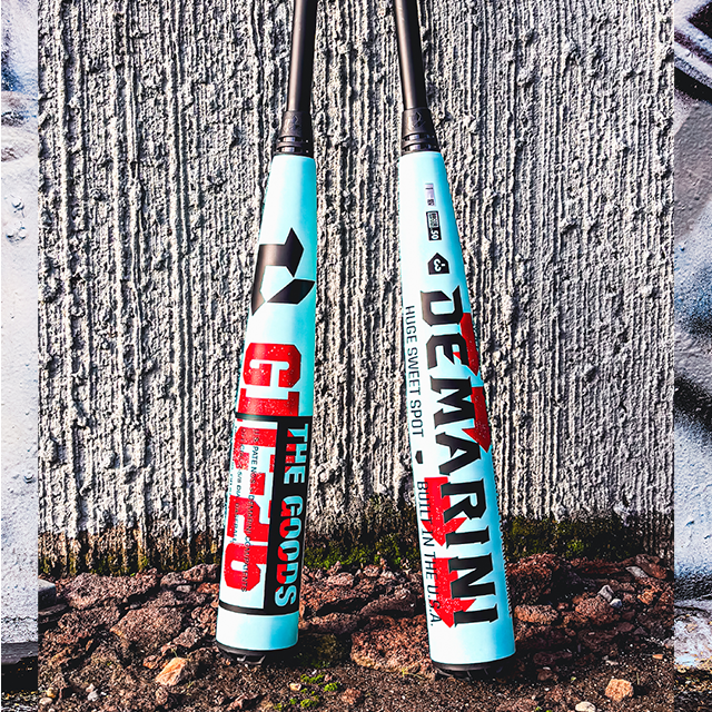 Two blue DeMarini baseball bats with bold red and black text lean against a textured concrete wall, standing on rough gravel and dirt.