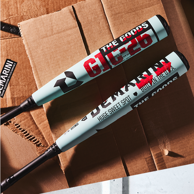 Two light blue DeMarini The Goods baseball bats with black handles rest on a ripped cardboard box. The bats have bold red and black graphics and slogans printed on them.