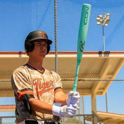 A baseball player in a camouflage Tigers jersey and helmet holds the 2026 Rawlings Icon (-3) BBCOR Baseball Bat: RBB6I3, standing on a field with a chain-link fence and stadium lights under a clear blue sky.