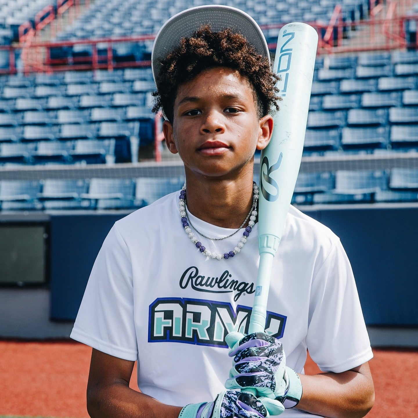 A young baseball player in a white “Rawlings ARMY” shirt and cap holds a bat while standing on a baseball field with empty stands in the background.