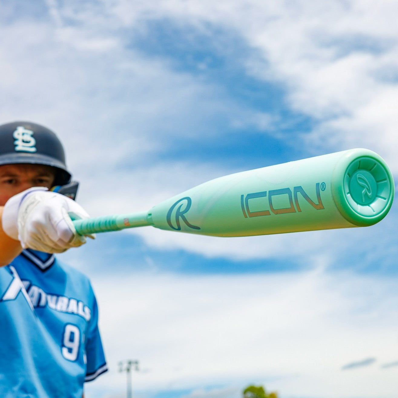 A baseball player in a blue jersey and black helmet holds the Rawlings 2026 Icon (-5) 2 3/4" USSSA Baseball Bat (RUT6I5) toward the camera, with a partly cloudy sky behind him.