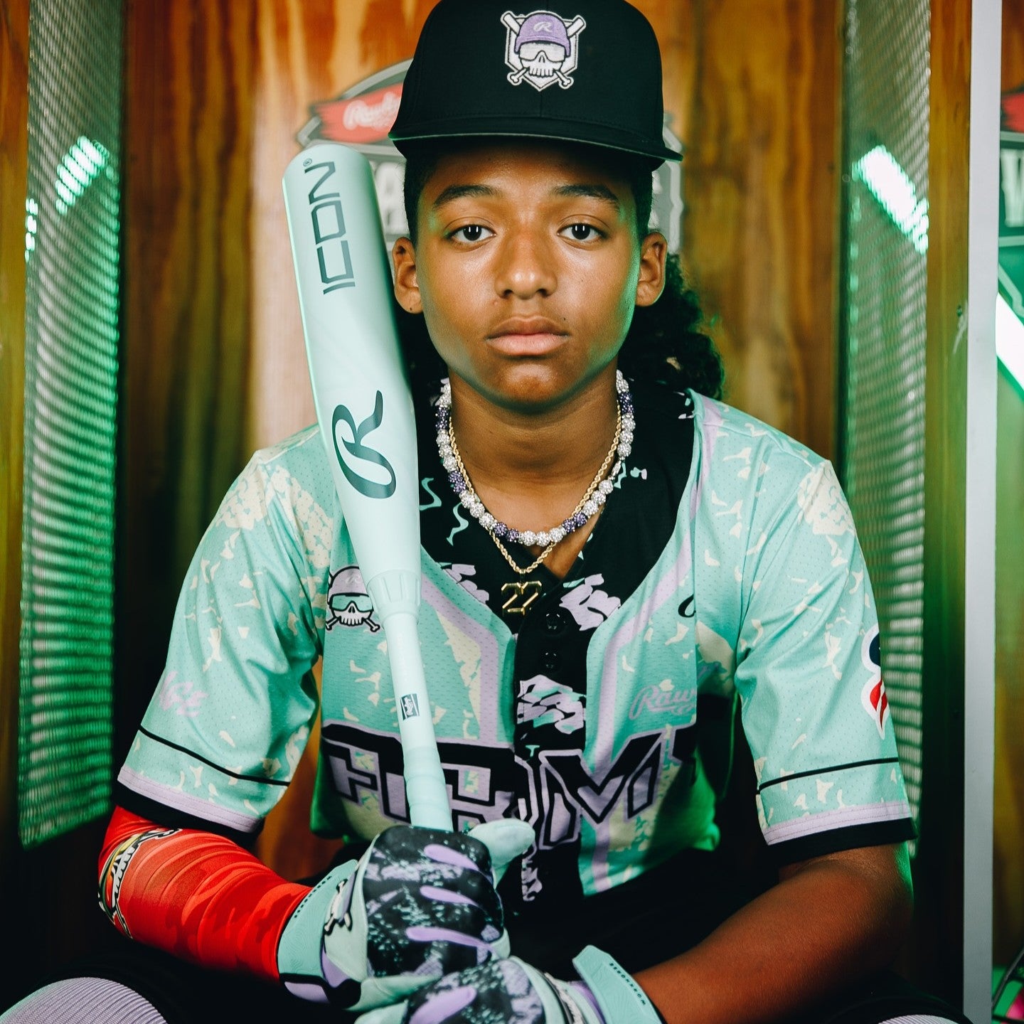 A young baseball player in a mint green uniform and black cap sits holding a bat upright. The player wears a necklace, arm sleeves, and batting gloves, and looks seriously at the camera with a colorful, lit background.