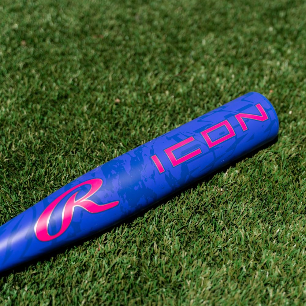 A blue 2027 Rawlings Icon (-5) 2 3/4″ USSSA Baseball Bat (RUT7I5) featuring the word ICON and a pink stylized R logo, crafted from In/Tense Carbon Composite in a two-piece composite design, rests on green grass.