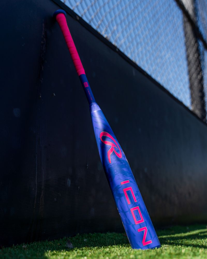 A Rawlings 2027 Icon (-5) 2 3/4″ USSSA Baseball Bat (model RUT7I5), featuring advanced two-piece composite construction in blue and pink, stands upright against a black fence on green artificial turf near a chain-link fence.