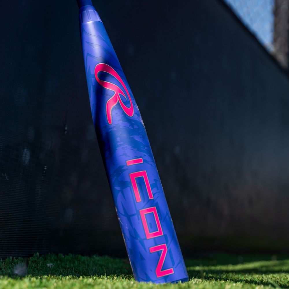 A blue and purple 2027 Rawlings Icon (-5) 2 3/4″ USSSA Baseball Bat (RUT7I5), crafted with In/Tense Carbon Composite, stands upright on artificial turf near a black fence, featuring bright pink lettering and sunlight casting shadows.