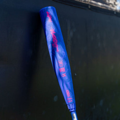 A blue Rawlings 2027 Icon (-5) 2 3/4″ USSSA Baseball Bat (model RUT7I5), adorned with pink writing and graphics, is propped against a dark outdoor wall. This bat showcases a two-piece composite design with In/Tense Carbon Composite technology, making it a standout choice for players.