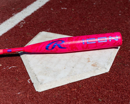 A vibrant pink Rawlings 2027 ICON (-10) 2 5/8″ USA Baseball Bat (RUS7I10) is positioned diagonally across a weathered home plate, set against the backdrop of a red turf field, highlighting the bat’s bold design.