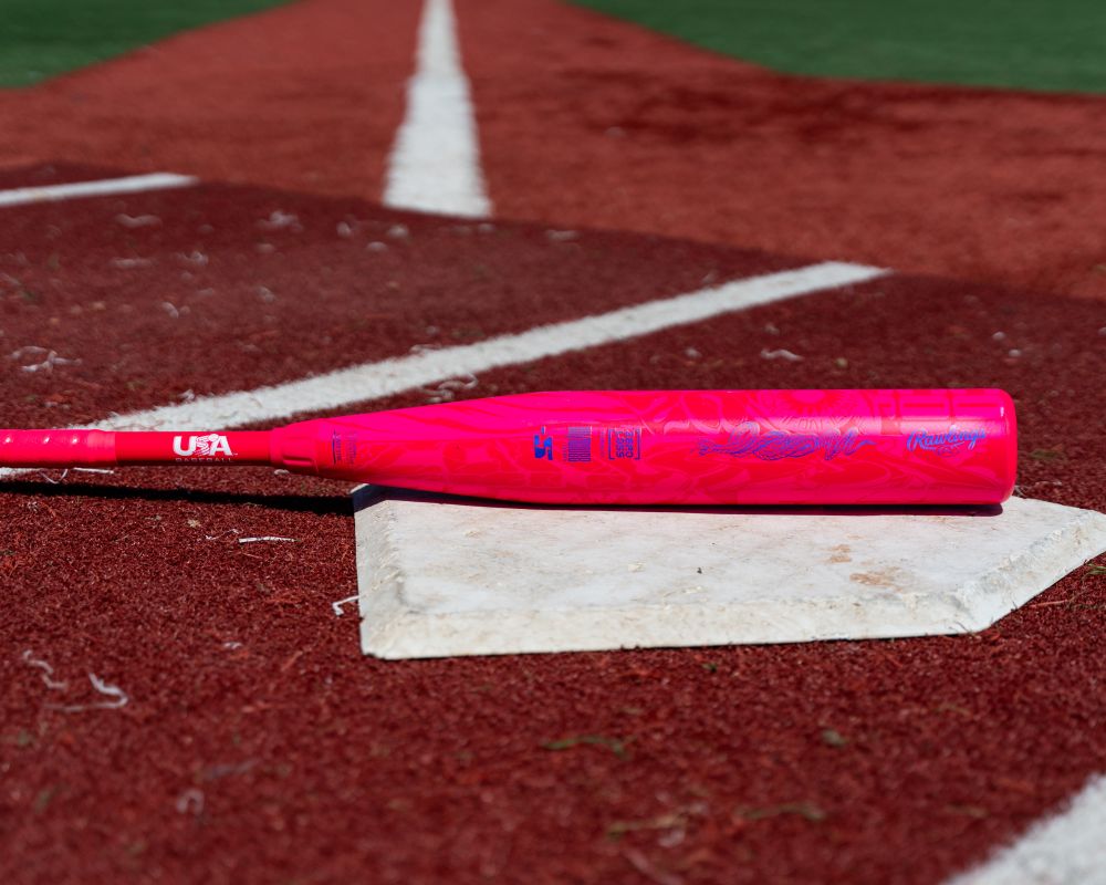 A Rawlings ICON (-10) 2 5/8″ USA Baseball Bat (RUS7I10) rests on home plate of a baseball field, with artificial turf and white baselines visible in the background.