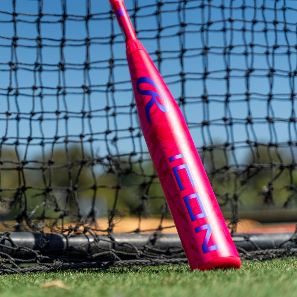 A vibrant pink 2027 Rawlings ICON (-10) 2 5/8″ USA Baseball Bat (RUS7I10), featuring bold blue “ICON” lettering, rests against a black net on a sunlit field. Lush grass and blurred trees in the background highlight this eye-catching Rawlings bat in action.