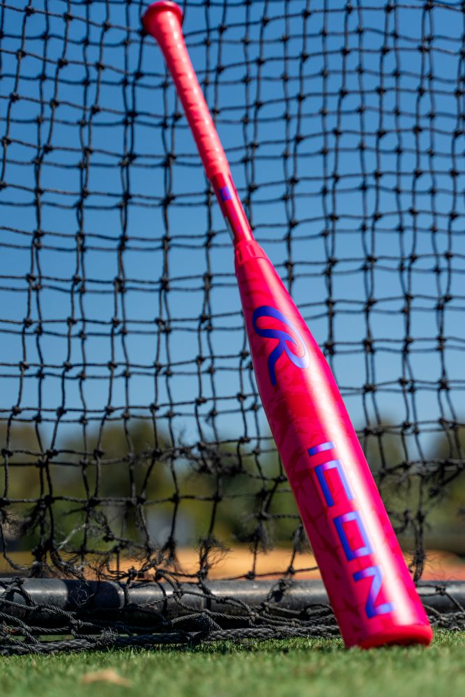 A vibrant pink 2027 Rawlings ICON (-5) 2 5/8″ USA Baseball Bat (model RUS7I5), featuring bold blue ICON lettering, rests against a black net on a grassy field beneath a clear blue sky and tall trees. This eye-catching composite bat from Rawlings is ideal for young players eager to make an impression on the diamond.