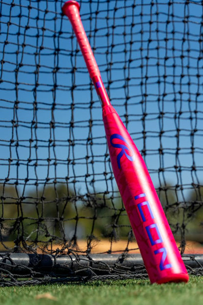 A vibrant pink 2027 Rawlings ICON (-5) 2 5/8″ USA Baseball Bat (model RUS7I5), featuring bold blue ICON lettering, rests against a black net on a grassy field beneath a clear blue sky and tall trees. This eye-catching composite bat from Rawlings is ideal for young players eager to make an impression on the diamond.