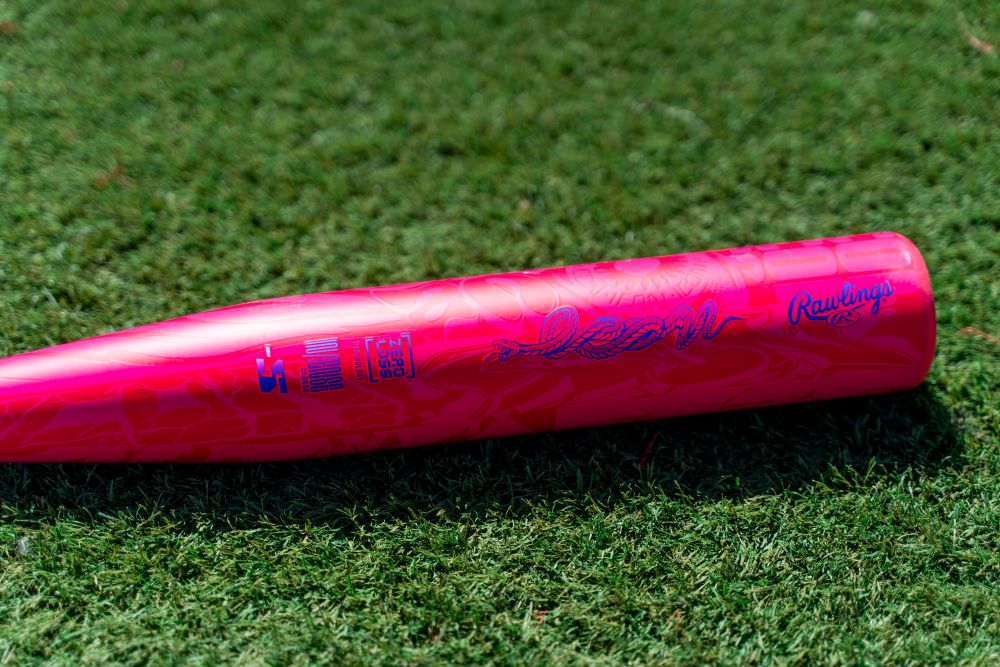 A vibrant pink Rawlings ICON (-5) 2 5/8″ USA Baseball Bat (RUS7I5) featuring blue text and graphics rests on green grass.