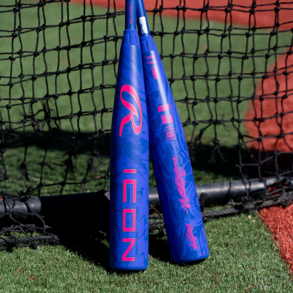 Two blue Rawlings 2027 Icon (-10) 2 3/4″ USSSA Baseball Bats (model RUT7I10), featuring pink lettering, stand upright on artificial turf near a black net and a red dirt area, suggesting a practice or game setting. These Rawlings bats include In/Tense Carbon Composite technology.