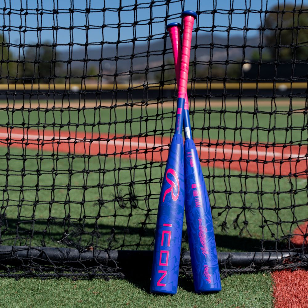 Two blue and pink 2027 Rawlings Icon (-8) 2 3/4″ USSSA Baseball Bats (model RUT7I8) lean against a black net on a baseball field, with green grass and a red dirt infield visible in the background.