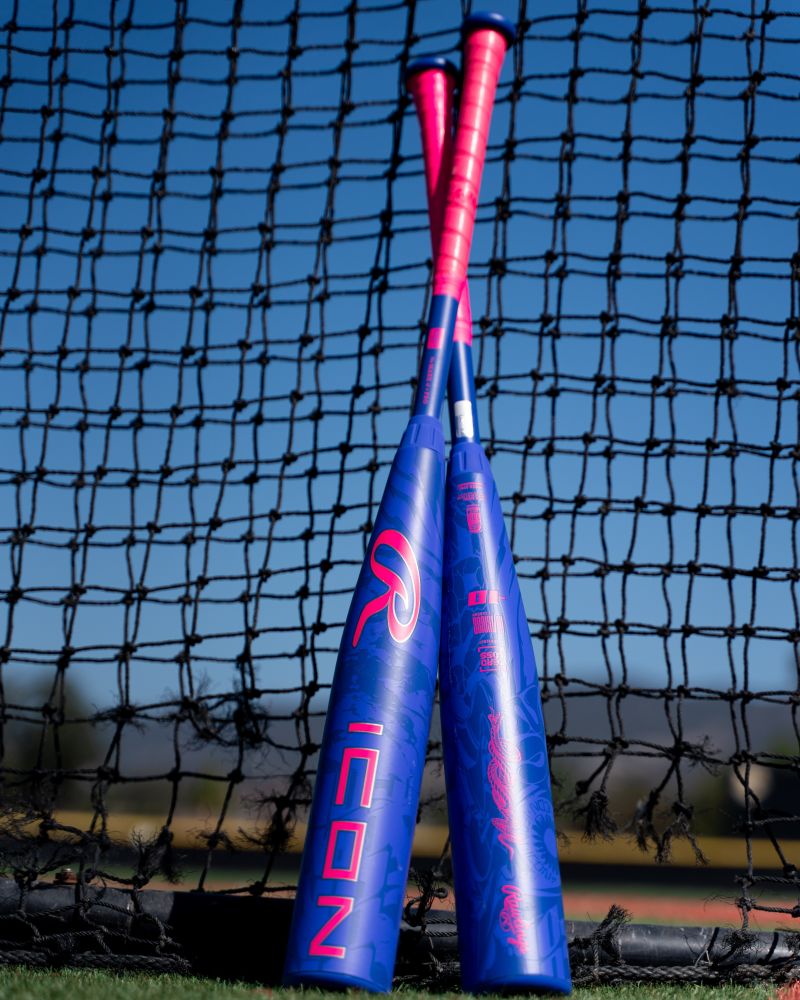 Two blue and pink baseball bats with bold graphics, including the 2027 Rawlings Icon (-5) 2 3/4″ USSSA Baseball Bat (RUT7I5) by Rawlings—featuring a two-piece composite design with In/Tense Carbon Composite technology—lean against each other in front of a black net on a sunny outdoor field.