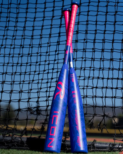 Two blue and pink baseball bats with bold graphics, including the 2027 Rawlings Icon (-5) 2 3/4″ USSSA Baseball Bat (RUT7I5) by Rawlings—featuring a two-piece composite design with In/Tense Carbon Composite technology—lean against each other in front of a black net on a sunny outdoor field.