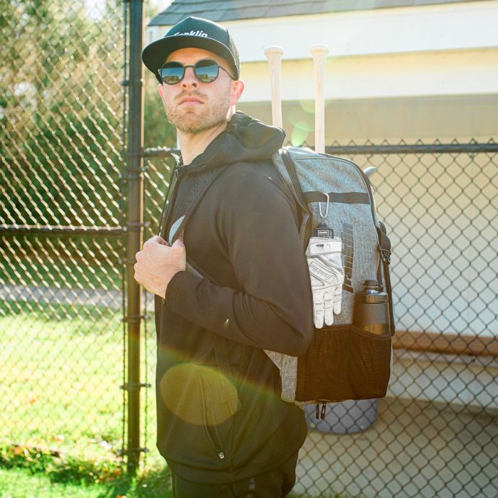 A man wearing sunglasses, a black hoodie, and a cap stands by a chain-link fence with the Franklin MLB Glove Aerator: 2357 sports backpack from Franklin, which fits two bats, batting gloves, a water bottle, and offers moisture control.