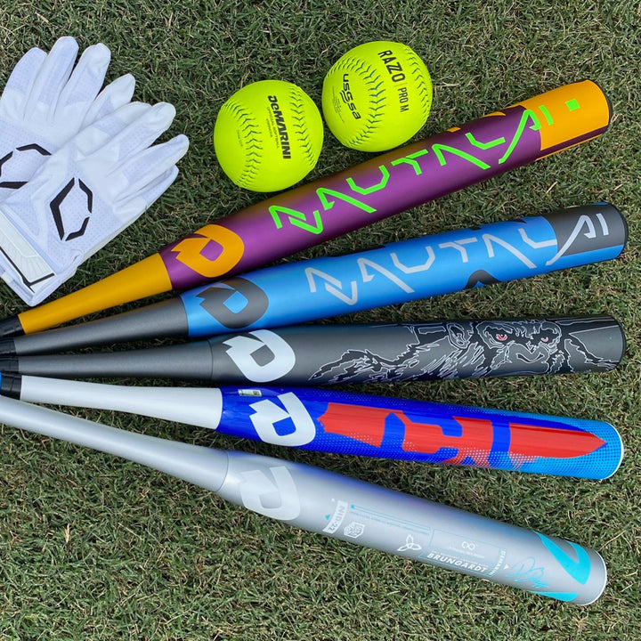 Five colorful baseball bats, two yellow softballs, and a pair of white batting gloves are arranged on green grass.
