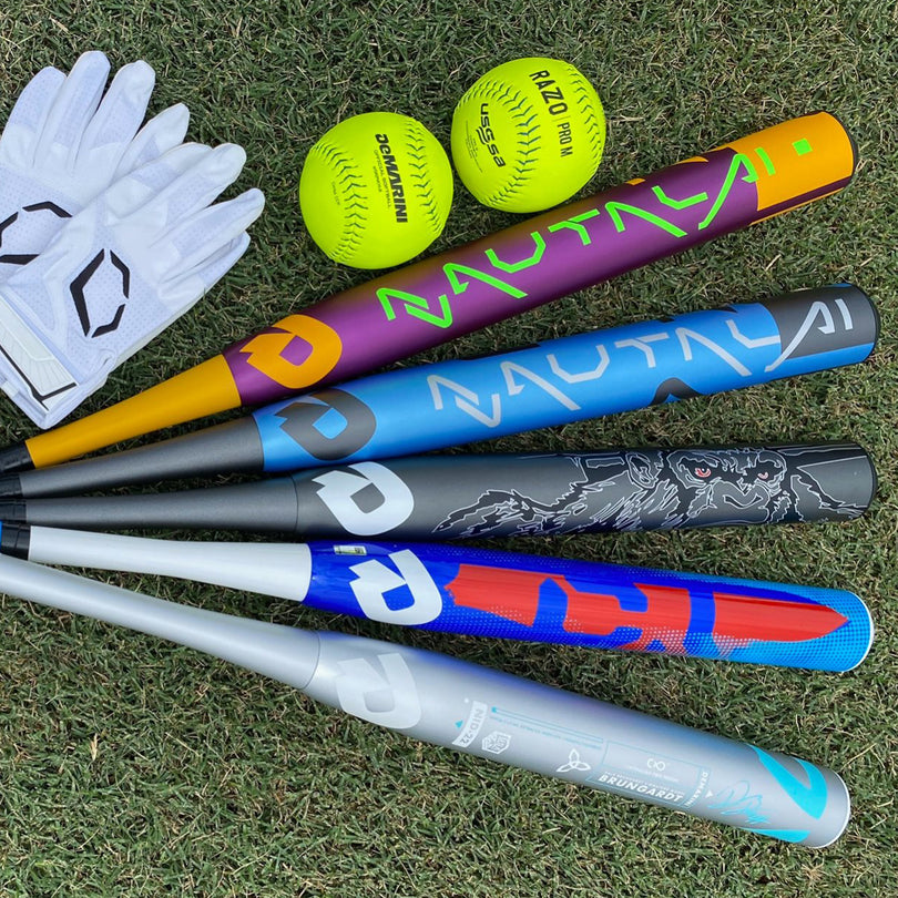 Five colorful baseball bats, two yellow softballs, and a pair of white batting gloves are arranged on green grass.